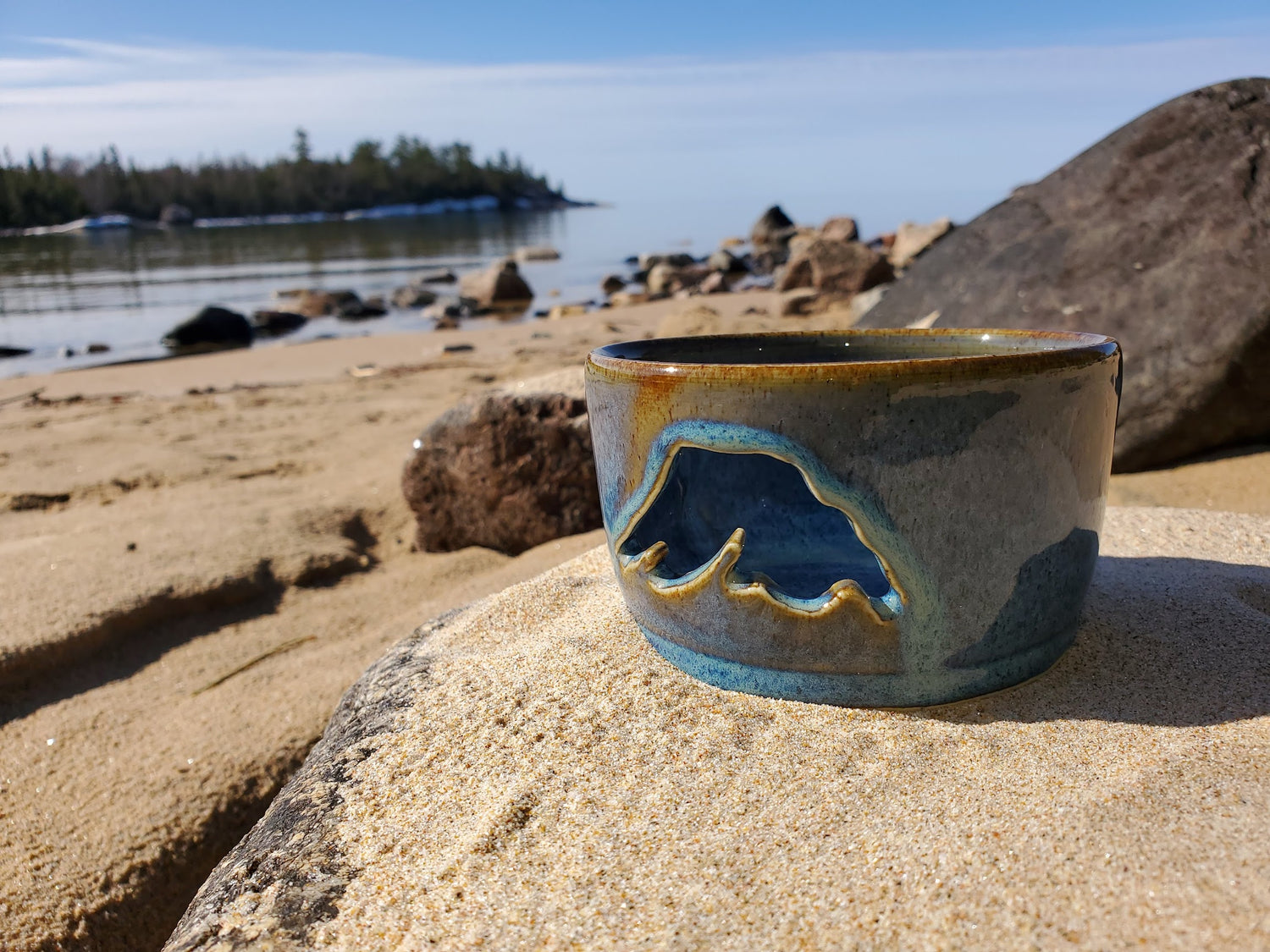 A brown and blue hand made pottery gift that looks like a candle holder sitting atop a rock on the beach of Lake Superior near Wawa, Ontario.
