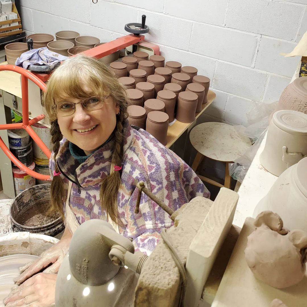 A photo of Cindy the maker and founder of Green Cabin pottery sitting in her pottery studio in Wawa, Ontario. Cindy is in the process of hand throwing pottery. Behind her are hand made mugs and hand made coffee mugs. Cindy is smiling in the photo, wearing a colourful sweater and her hair is in braids.