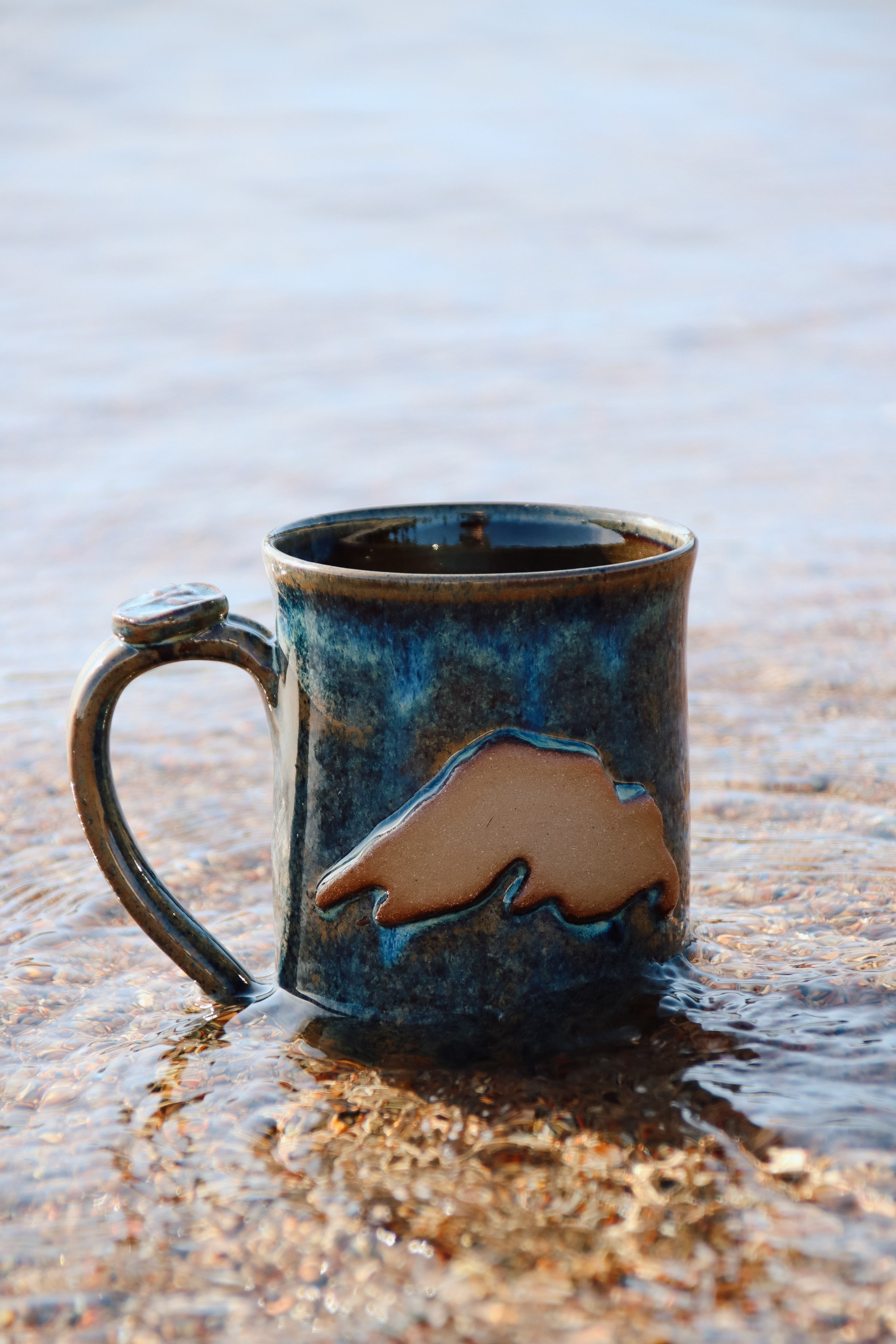 A blue hand made pottery coffee mug with a tan design of Lake Superior sitting on the beach, in the shallow water of Lake Superior near Wawa, Ontario.