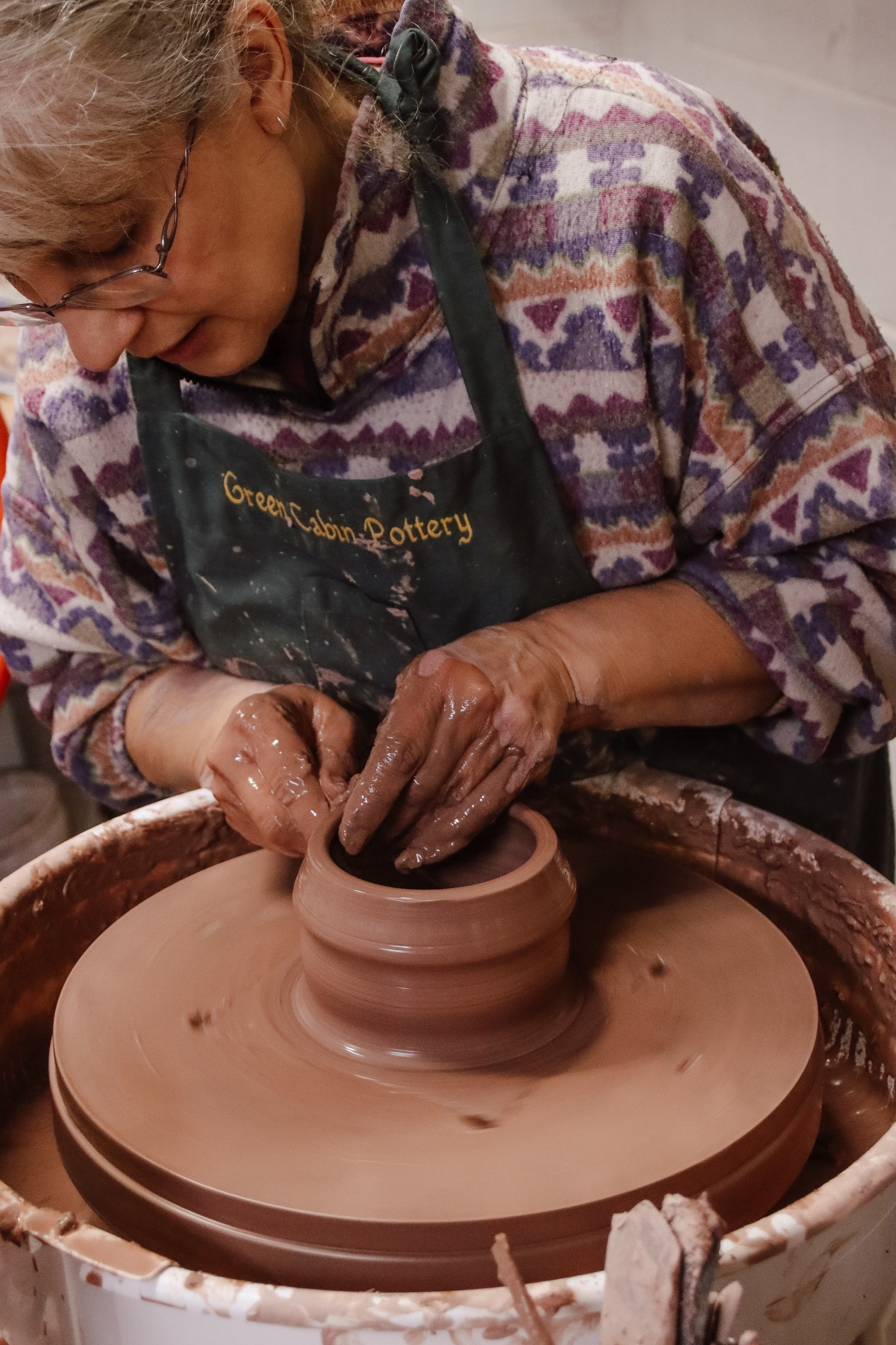 Cindy, maker and founder of Green Cabin Pottery is hand throwing clay on her pottery wheel creating a hand made coffee mug. Cindy is wearing a colourful sweater and an apron.