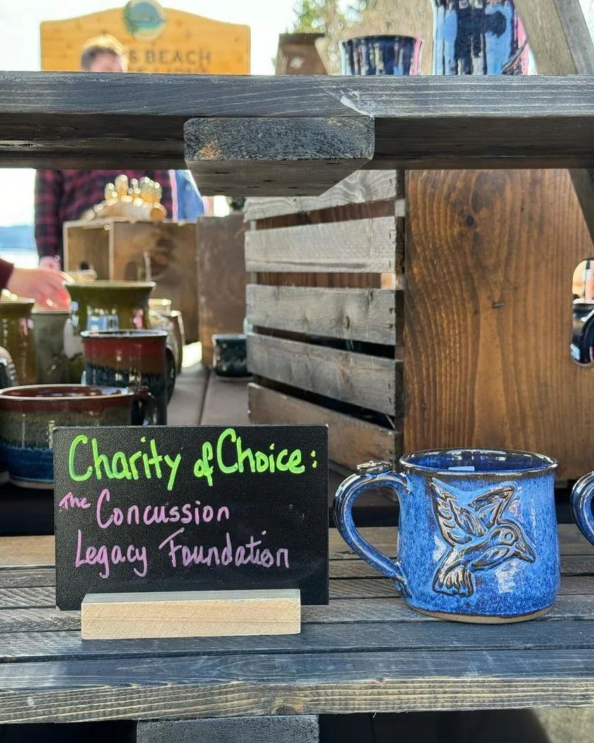 A blue hand made pottery mug sitting at a market in Wawa, Ontario beside a sign that says "Charity of Choice - the Concussion Legacy Foundation".