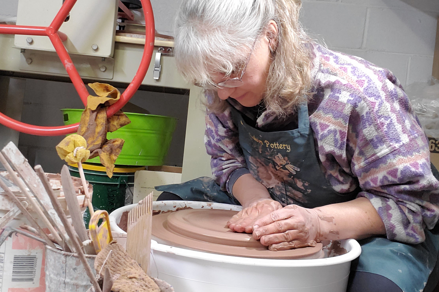 Cindy, maker and founder of Green Cabin Pottery is hand throwing clay on her pottery wheel creating a hand made pottery gift. Cindy is wearing a colourful sweater and an apron.