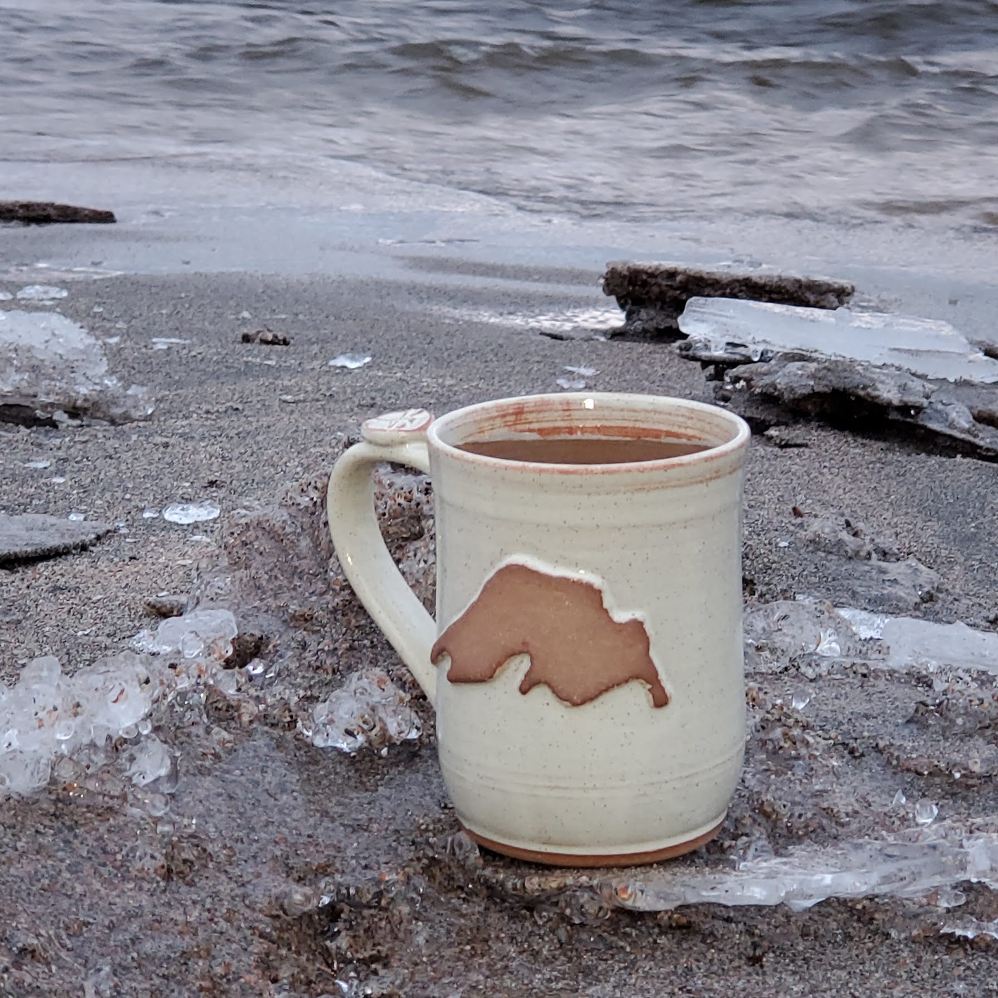 A white hand made coffee mug sits on the shore of Lake Superior near Wawa Ontario. The hand made mug features a brown design of Lake Superior. The photo is taken in the winter and there is ice on the sand.