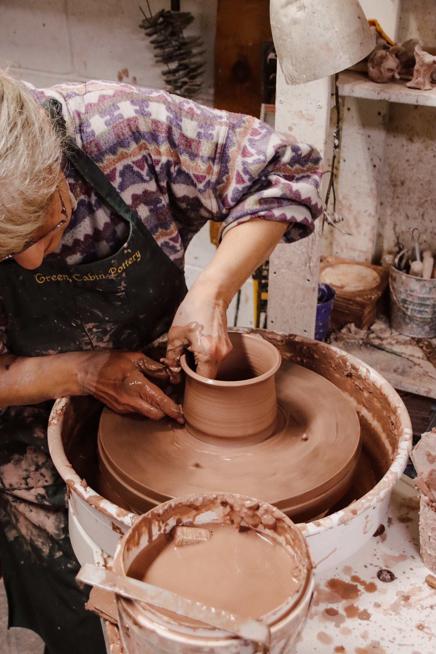 Cindy, maker and founder of Green Cabin Pottery is hand throwing clay on her pottery wheel creating a hand made coffee mug. Cindy is wearing a colourful sweater and an apron.