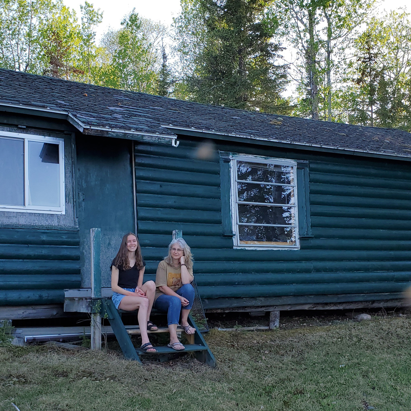Cindy, the maker and founder of Green Cabin Pottery and her daughter sit outside of a green cabin near Wawa, Ontario. This cabin was the inspiration for the name of her company. They are smiling and in summer clothes.